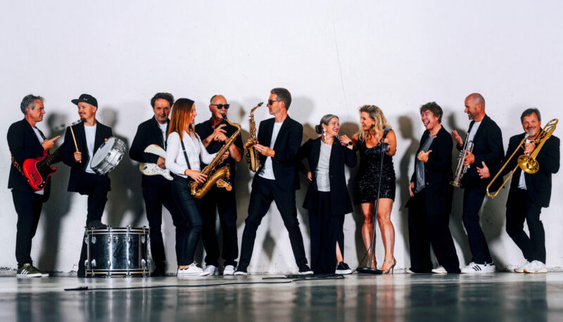 Live-Band mit Bläser-Section und drei Sängerinnen spielt Disco-Soul-Funk auf einer Bühne.