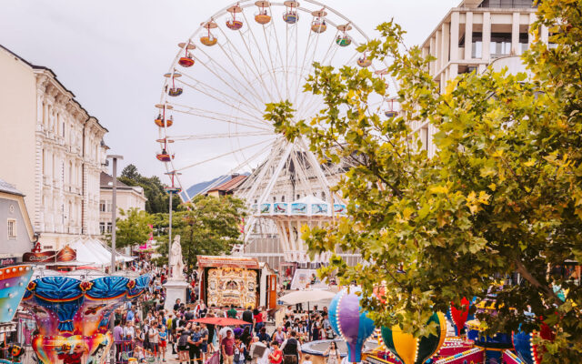 Besucherinnen und Besucher feiern beim Villacher Kirchtag, im Hintergrund ein Zug als Symbol der Anreise mit der Westbahn.